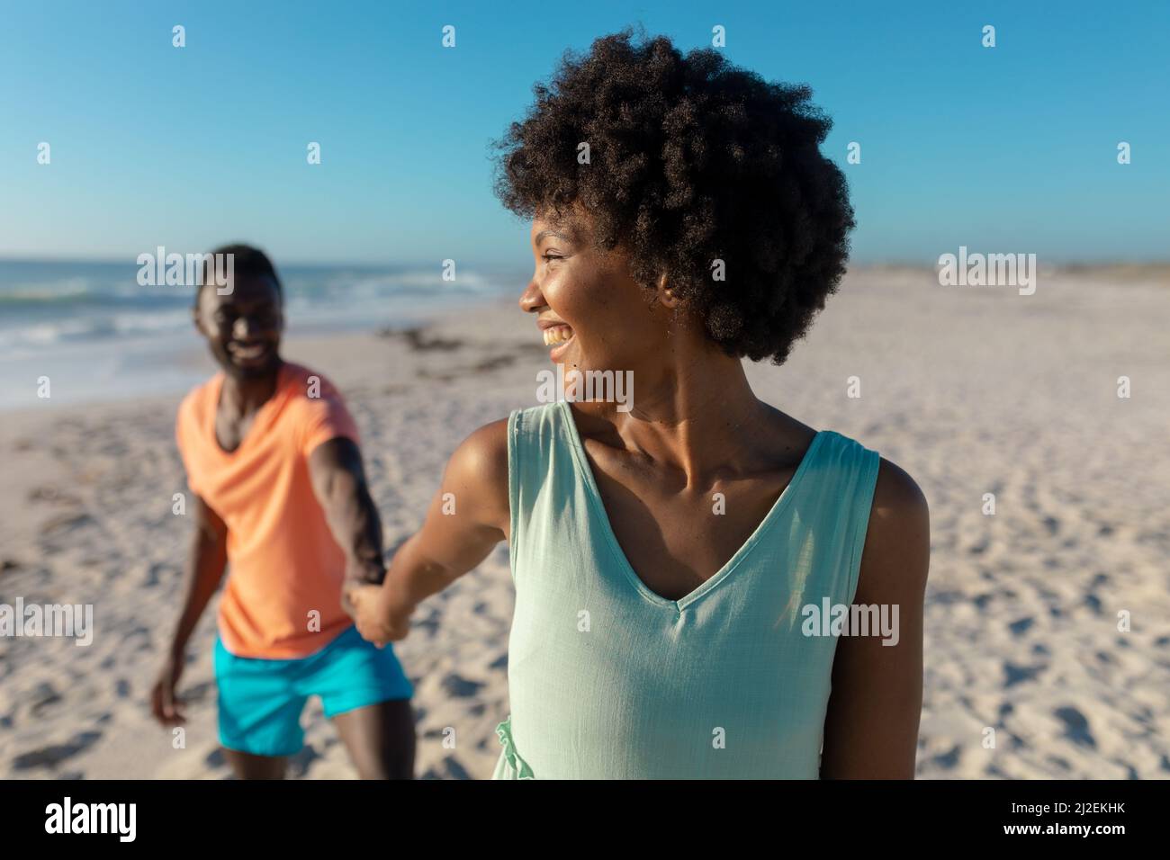 Smiling african american woman looking over shoulder while leading boyfriend at beach on sunny day Stock Photo