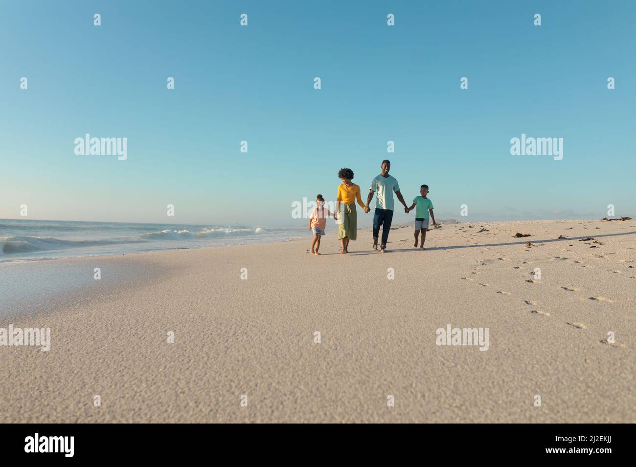 African american family holding hands while walking at beach against blue sky with copy space Stock Photo