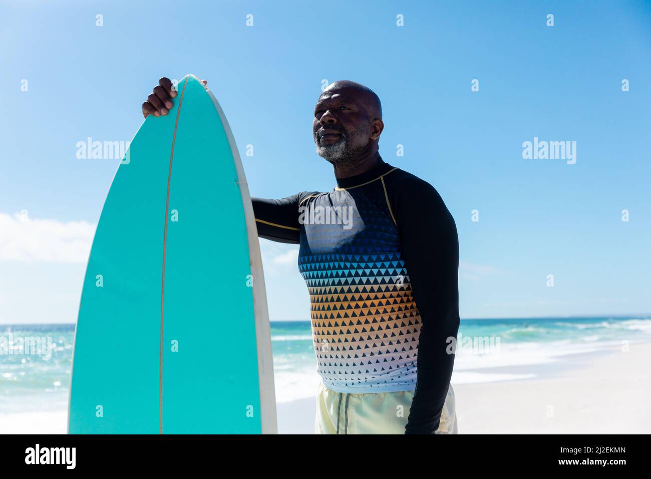 Retired senior african american man holding surfboard standing at beach against blue sky Stock Photo