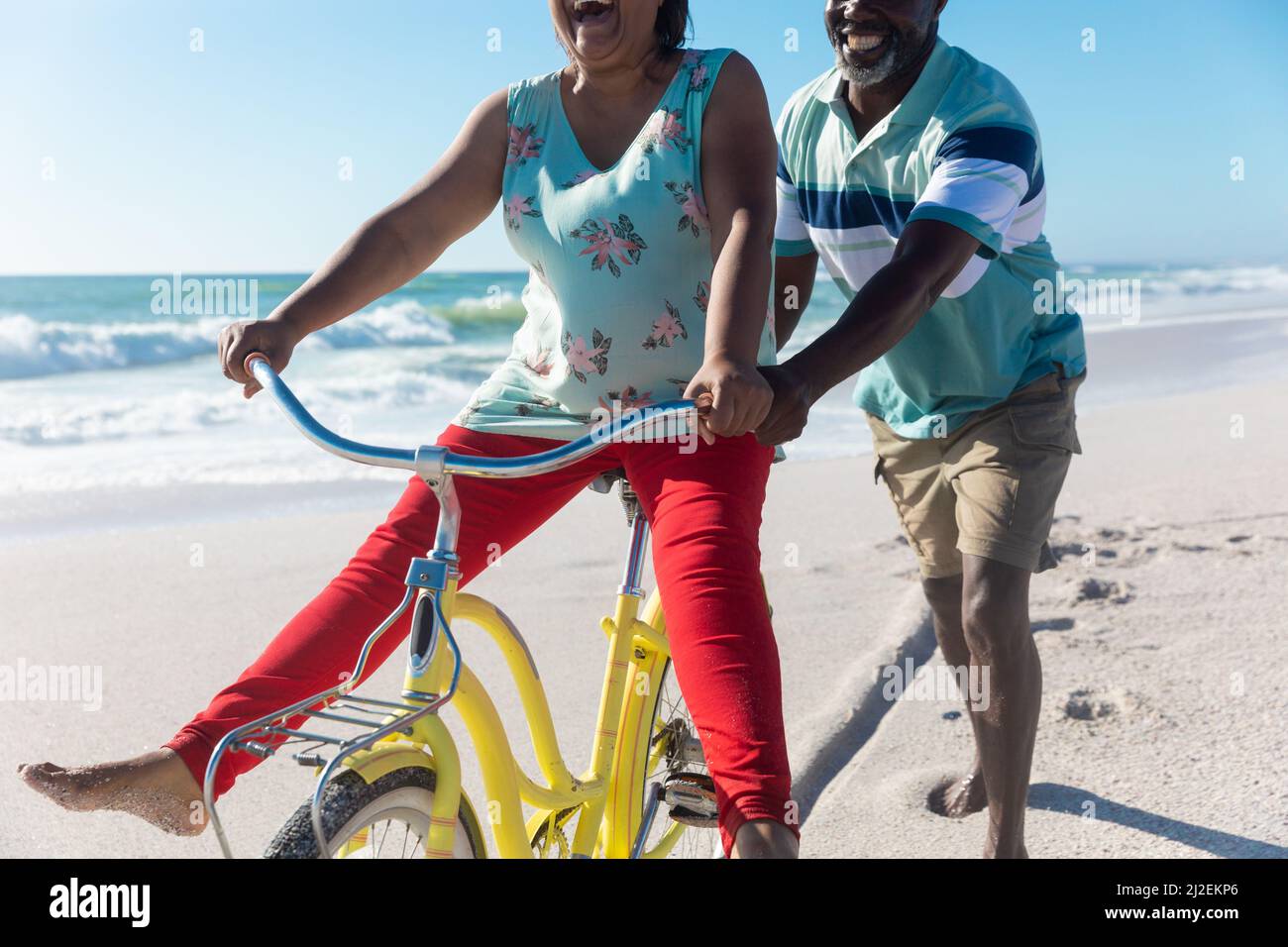 African american senior man assisting woman riding bicycle at beach on sunny day Stock Photo