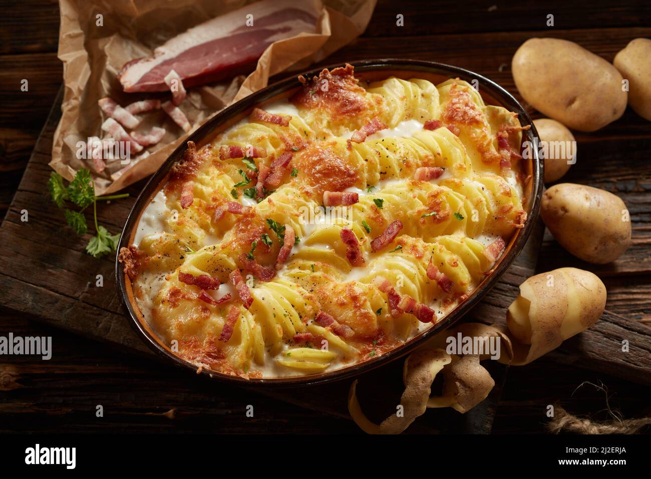 The top shot of a homemade traditional casserole with sliced potato and cheesy cream served in an oval pan with bacon pieces and parsley Stock Photo
