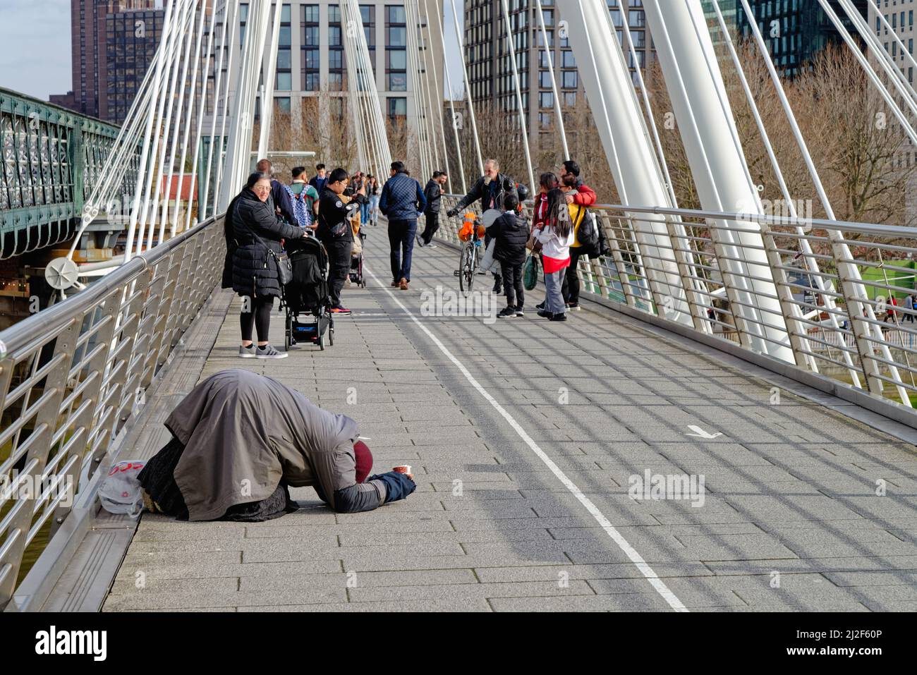 A lone beggar lying prostrate on the ground begging on the Golden Jubilee pedestrian bridge with tourists passing by, Charing Cross London England UK Stock Photo