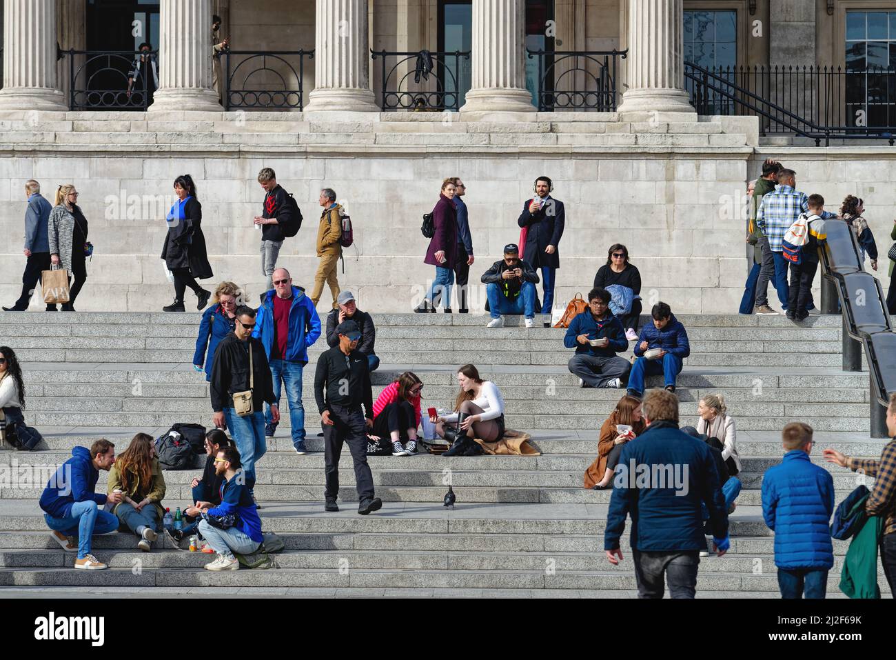 Crowds of tourists and pedestrian sitting on the steps outside the National Gallery in Trafalgar Square on a sunny spring day Central London England Stock Photo