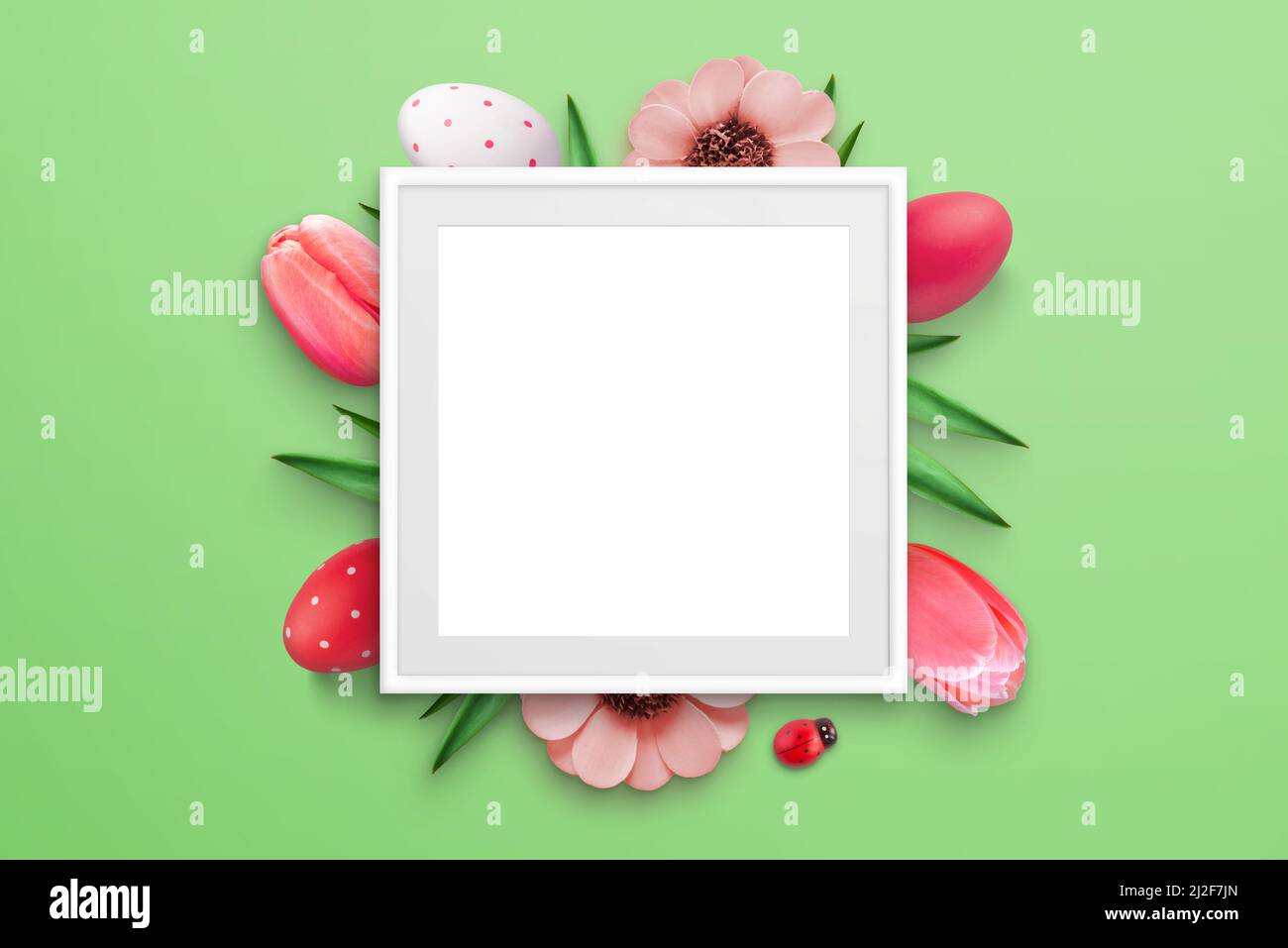 Blank picture frame surrounded by Easter eggs and flowers on green surface. Top view, flat lay composition Stock Photo