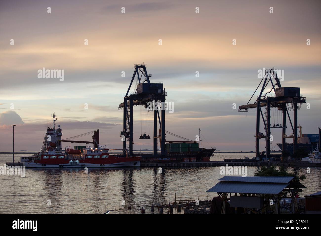 The evening sun sets over container cranes that stand at the port in Makassar, Indonesia. Stock Photo