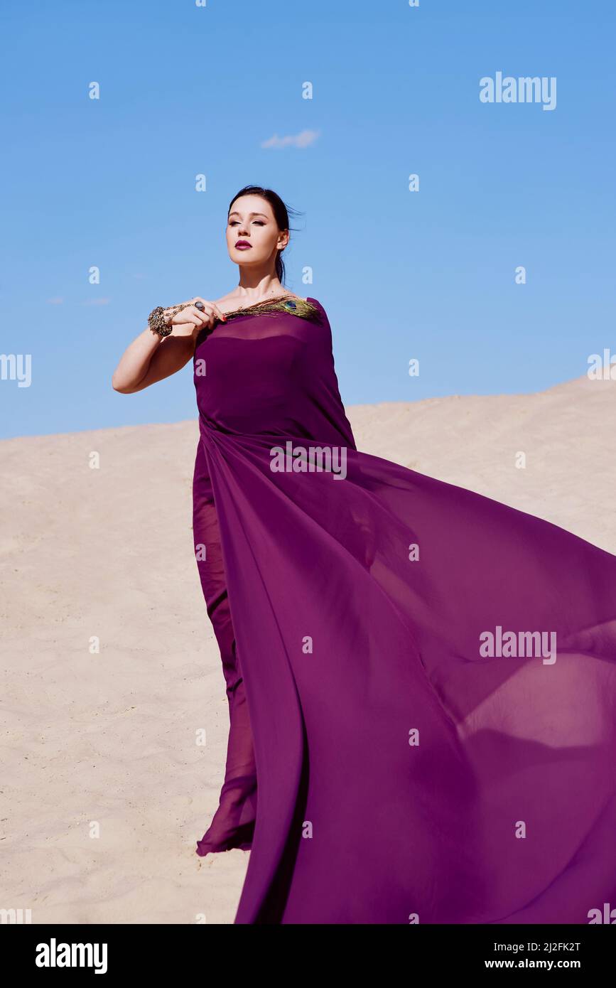Amazing beautiful brunette woman with the Peacock feather in purple fabric in the desert. Oriental, Indian, fashion, style concept Stock Photo