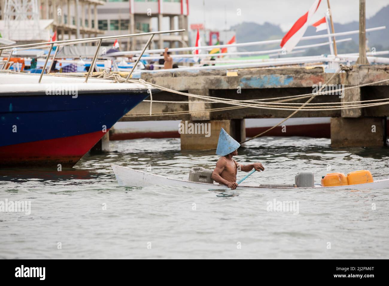 A man wearing a traditional conical hat steers a sinking canoe through the harbor and docks in Mamuju city, Sulawesi, Indonesia, Asia. Stock Photo