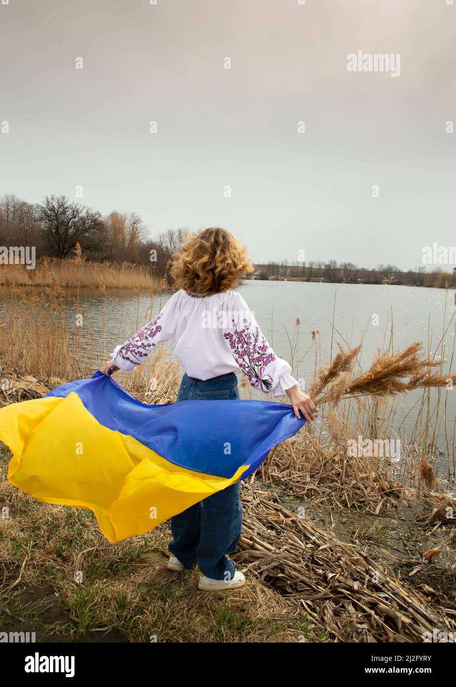 unrecognizable young woman stands with her back on the shore of a lake with a blue and yellow Ukrainian flag flying. pride of the nation, I am Ukraini Stock Photo