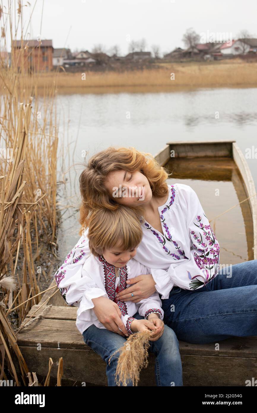 little boy and young beautiful woman in embroidered national clothes are sitting on shore of lake. Family, refugees, unity, support, patriotism. Ukrai Stock Photo