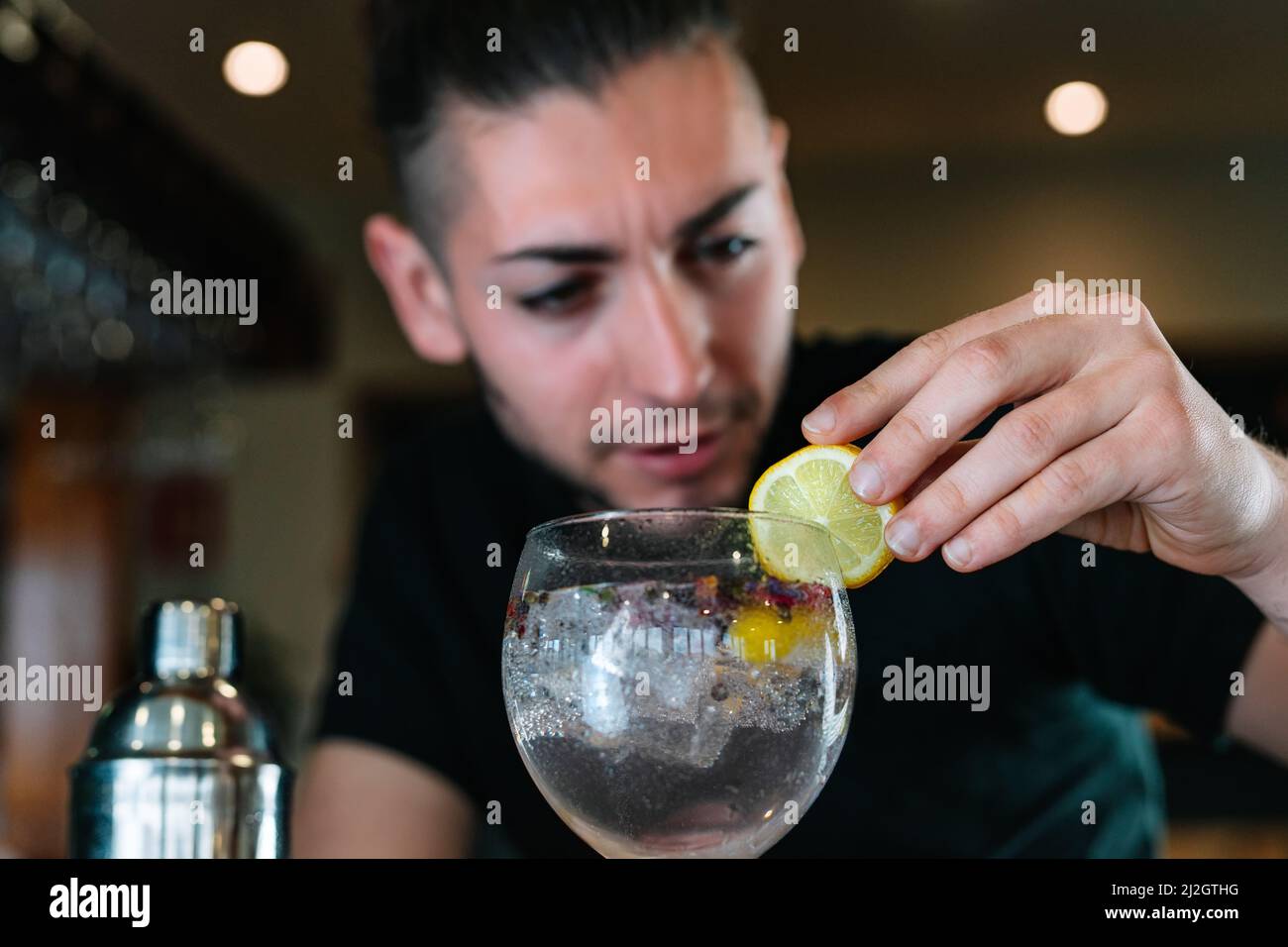 Young bartender garnishing with a slice of citrus fruit a cocktail in a crystal glass with ice. Stock Photo