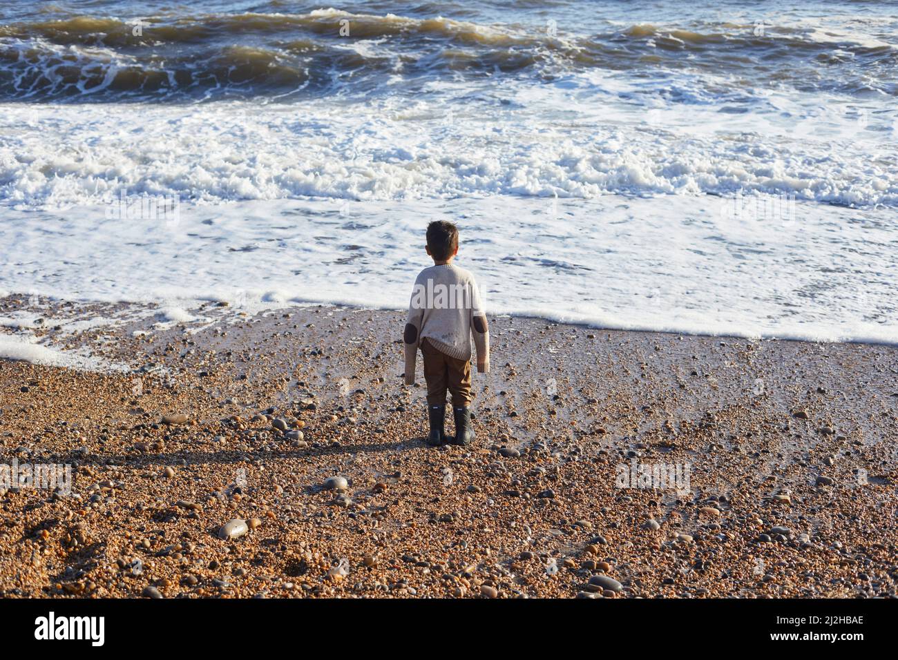 UK, Devon, Rear view of boy in oversized sweater looking at sea waves Stock Photo