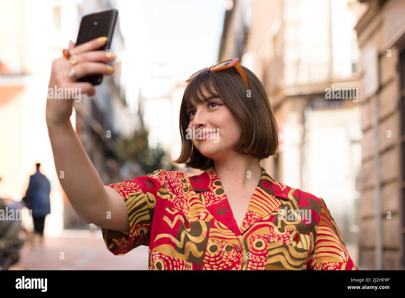 Smiling young caucasian woman with short hair taking a photo of herself with a mobile phone outdoors. Stock Photo