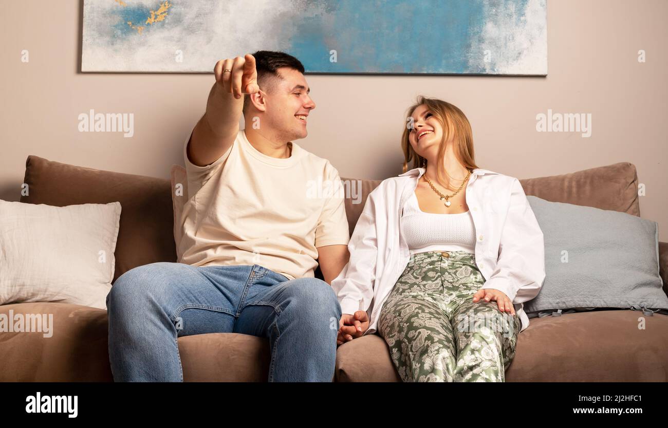 Minsk Belarus - April 02 2022 Happy couple sitting on sofa, laughing, watching TV and talking. Handsome man pointing at screen. Sincere, fun relations and love concept. Stock Photo