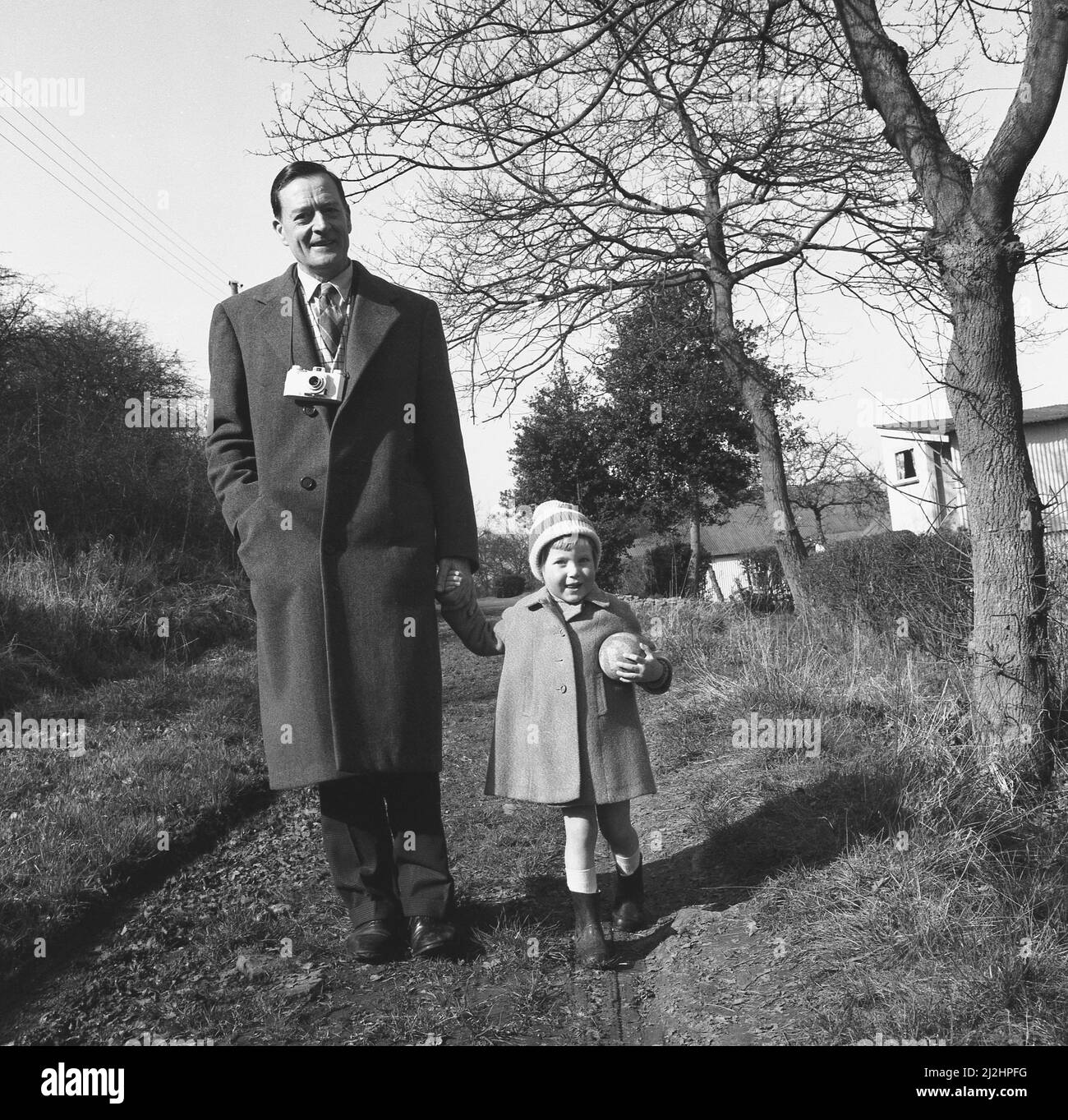 1950s, historical, a father out for a walk with young daughter, who in a coat and bobble hat is holding a small ball, England, UK. The man is wearing an overcoat and with a film camera hanging around his neck. Stock Photo