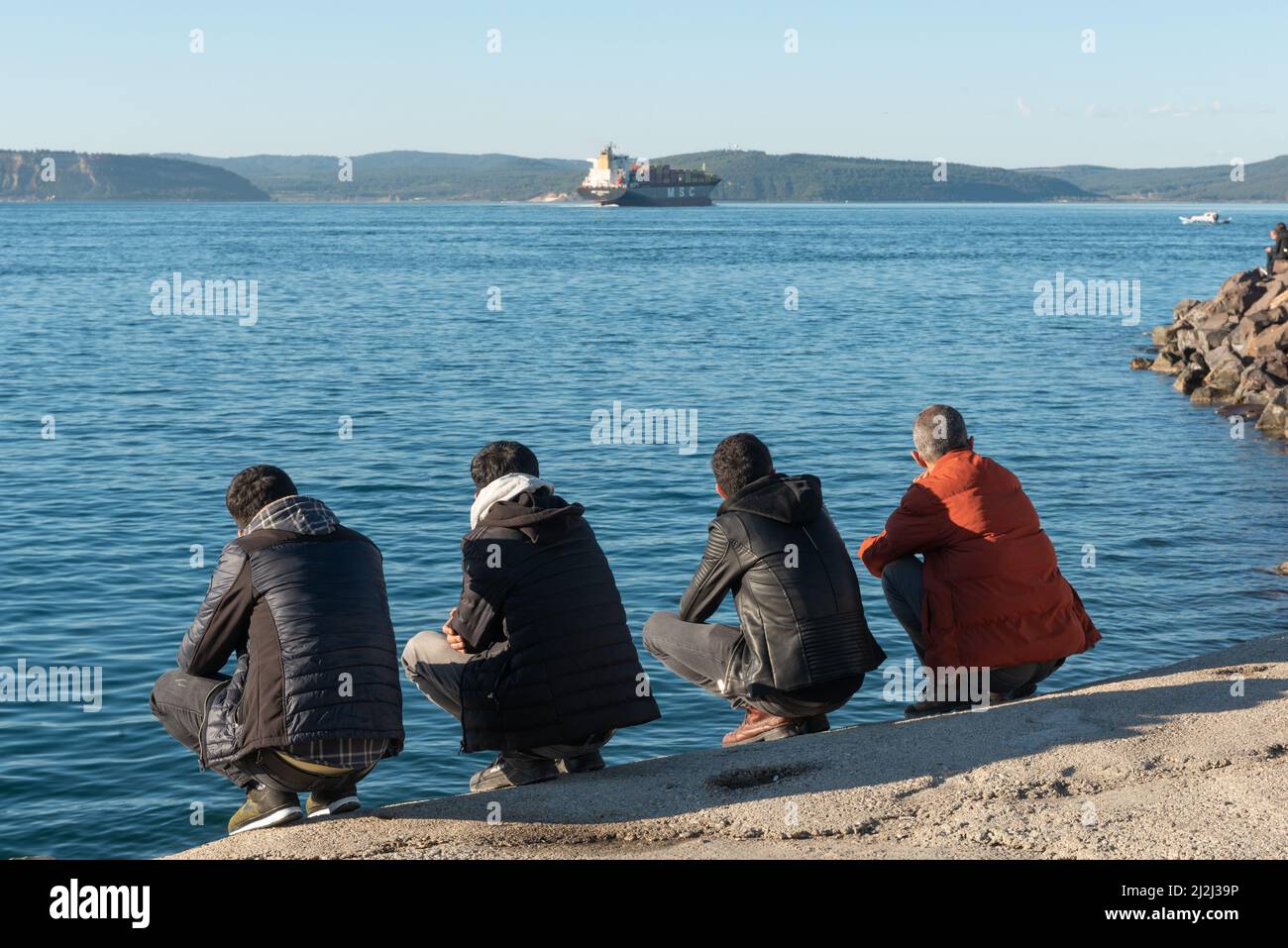 Canakkale, Turkey. February 18th 2022 Turkish men squatting beside the Dardanelles Straight opposite the Gallipoli Peninsular, the harbour of the port Stock Photo
