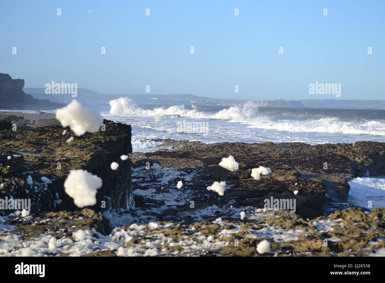 White Sea Foam Blowing Across Filey Brigg Rocks And Boulders - Windy Sunny Day - Rough North Sea - White Horses On North Sea - Yorkshire UK Stock Photo