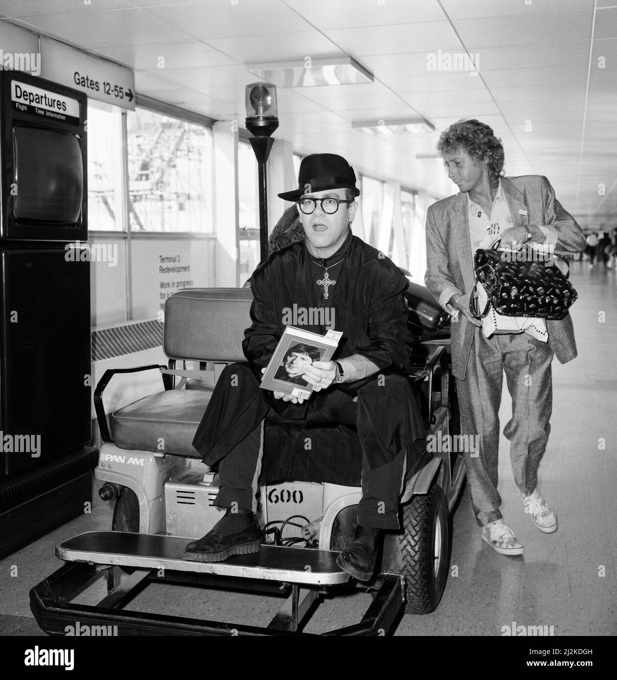 Elton John at Heathrow Airport, wearing a monk's black robe and a big gold cross, topped off with a black hat. He rode on a baggage trolley. He is on his way to California to collect a television award. London. 7th September 1987. Stock Photo