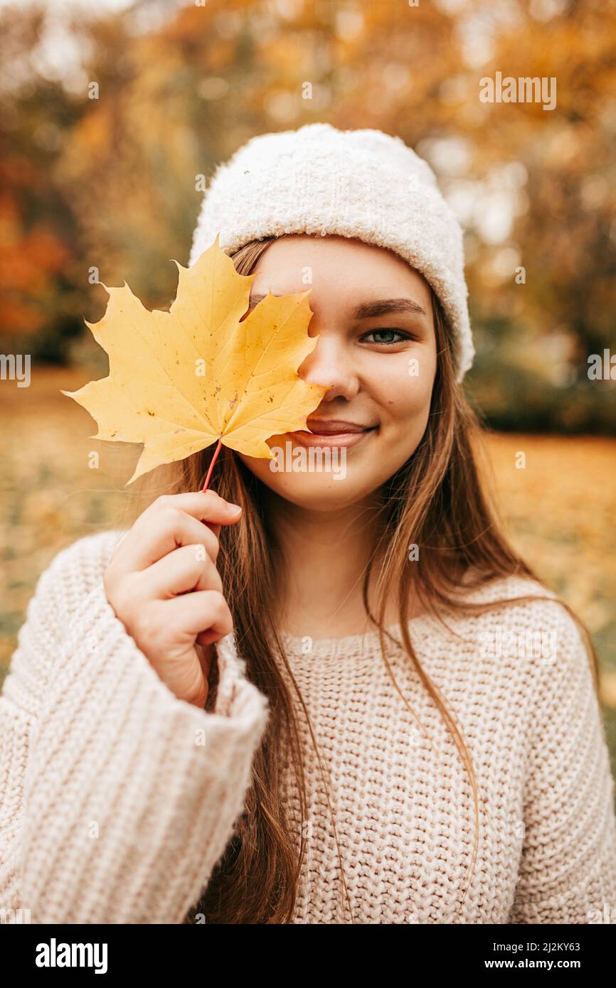 Vertical portrait of close up face attractive blond smiling woman in hat, hold golden maple leaf face, close eye. Autumn Stock Photo