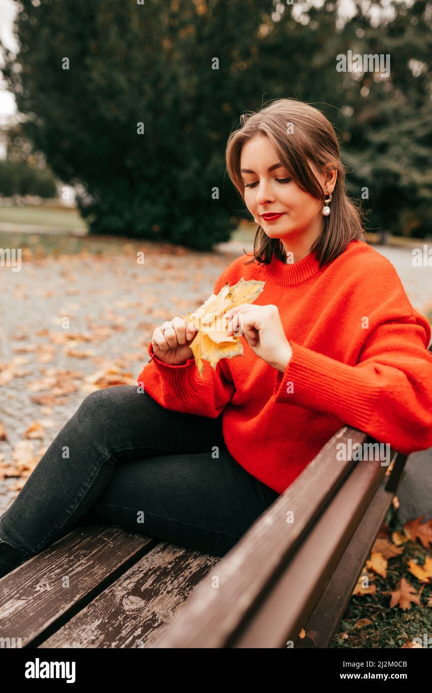 Vertical portrait of pretty thoughtful woman student, sitting on bench, hold bunch maple leaves. Park autumnal nature Stock Photo
