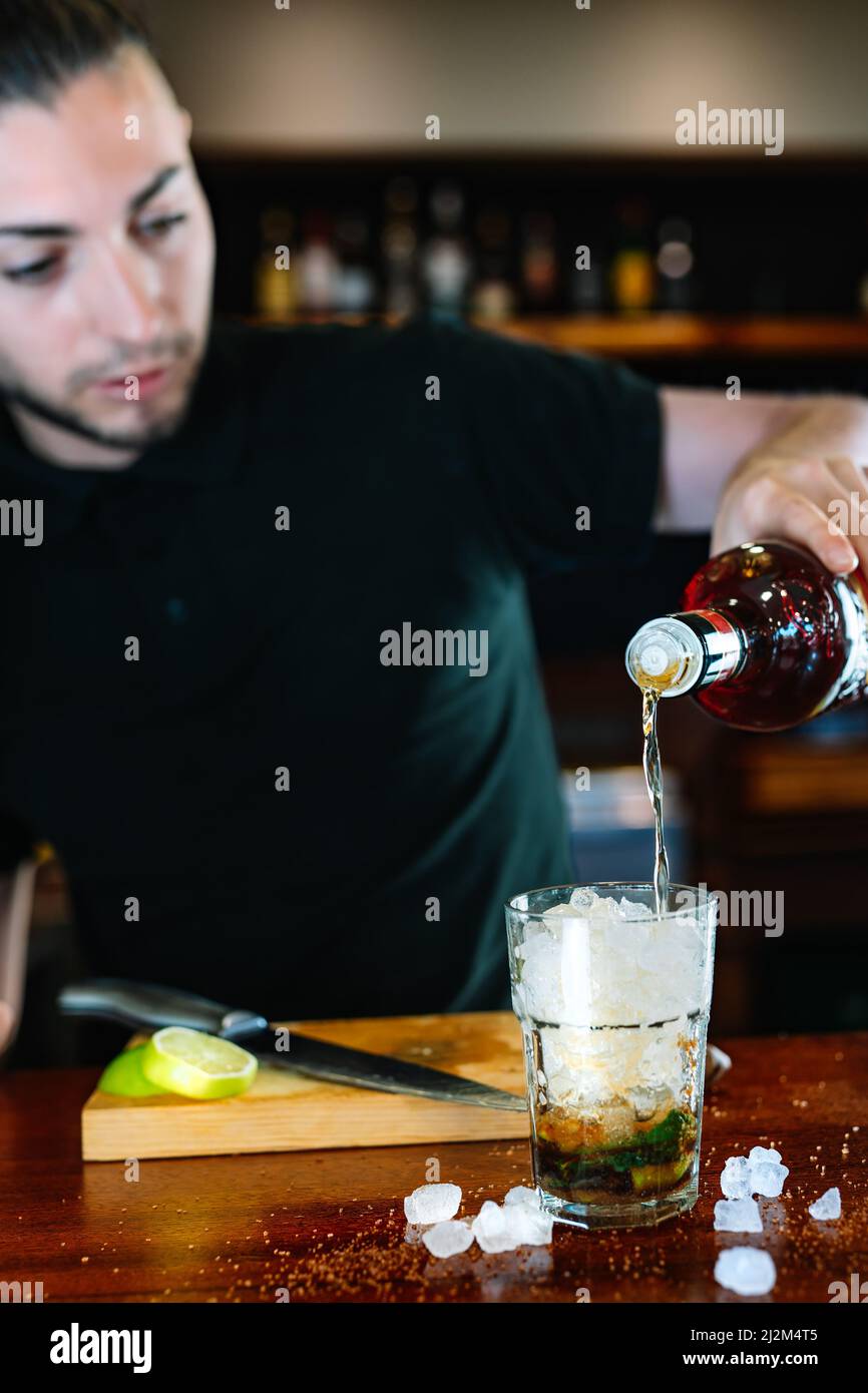 young waiter, pour liquor into a crystal glass filled with ice to prepare a Mojito. Vertical View Stock Photo