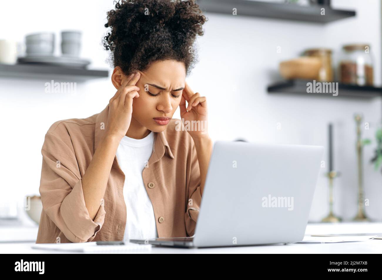 Tired african american girl overworked from working in a laptop, sitting at a table in the kitchen at home, closed her eyes, massages her temples, has a migraine, headache, tension, needs rest, break Stock Photo