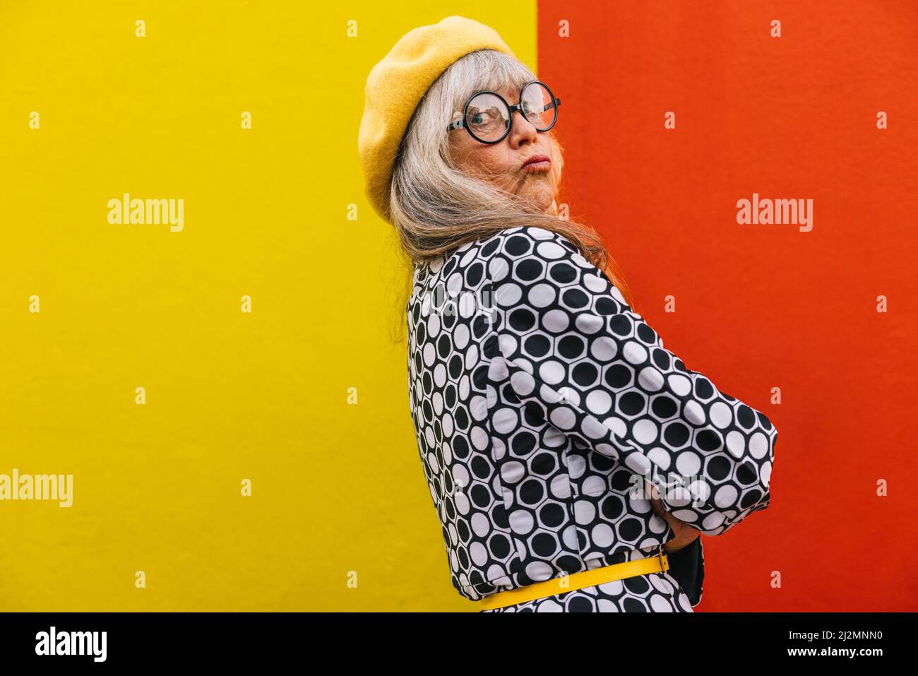 Funny senior woman looking at the camera with her lips pouted. Elderly woman standing against a red and yellow background with her arms crossed. Matur Stock Photo