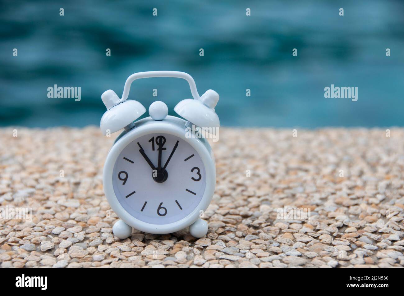 White alarm clock on marble floor with blurred swimming pool background. The clock set at 11 o'clock. Morning and deadline concept. Stock Photo