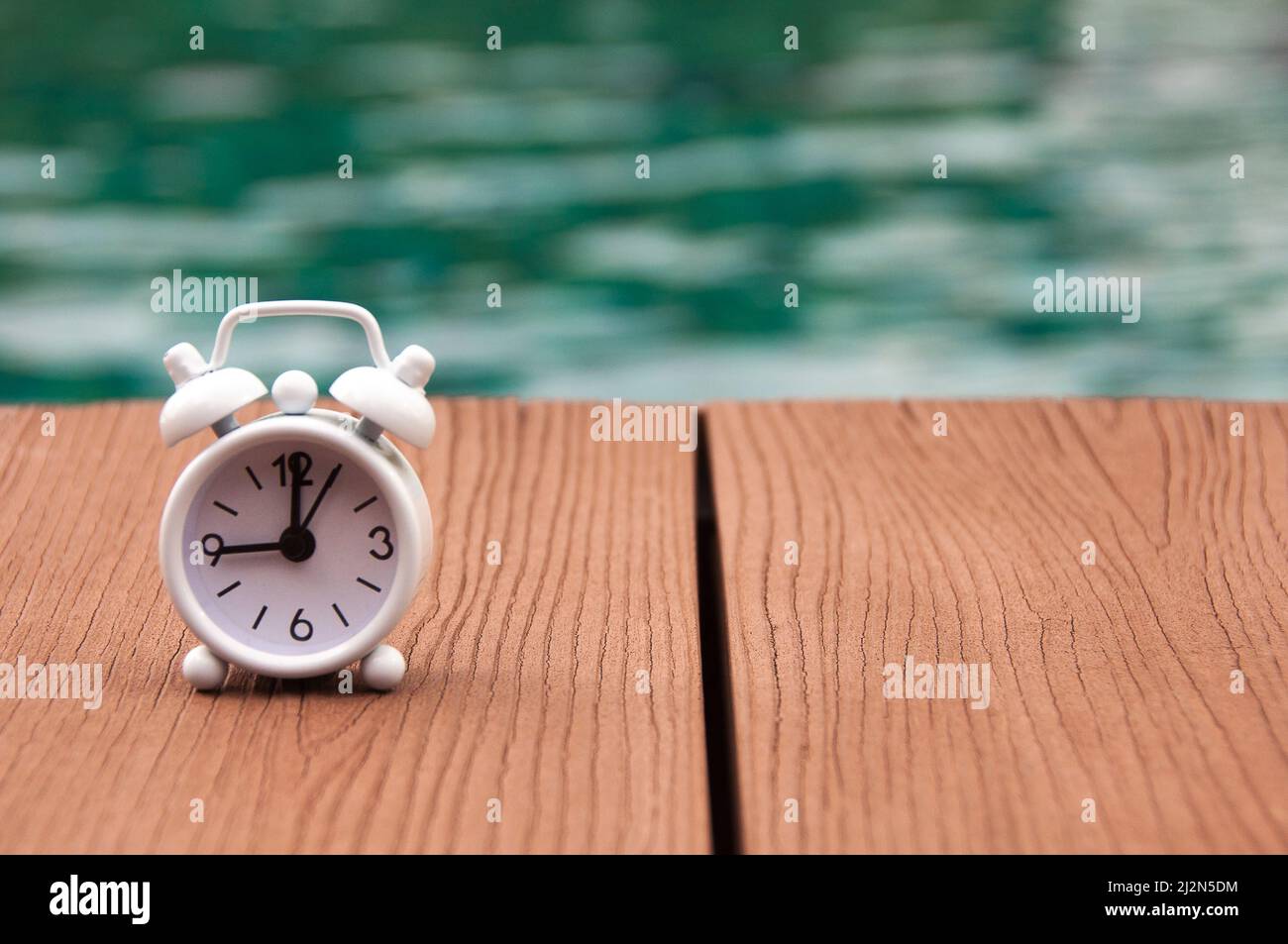 Alarm clock on wooden floor with blurred swimming pool background. The clock set at 9 o'clock. Stock Photo
