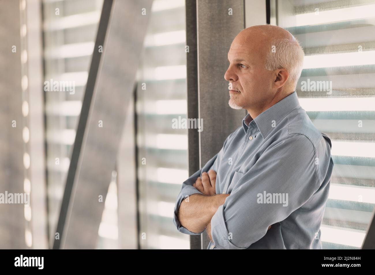 Middle-aged man leaning against a modern stained glass window of a company or futuristic building, looks out into the distance and sees even further a Stock Photo