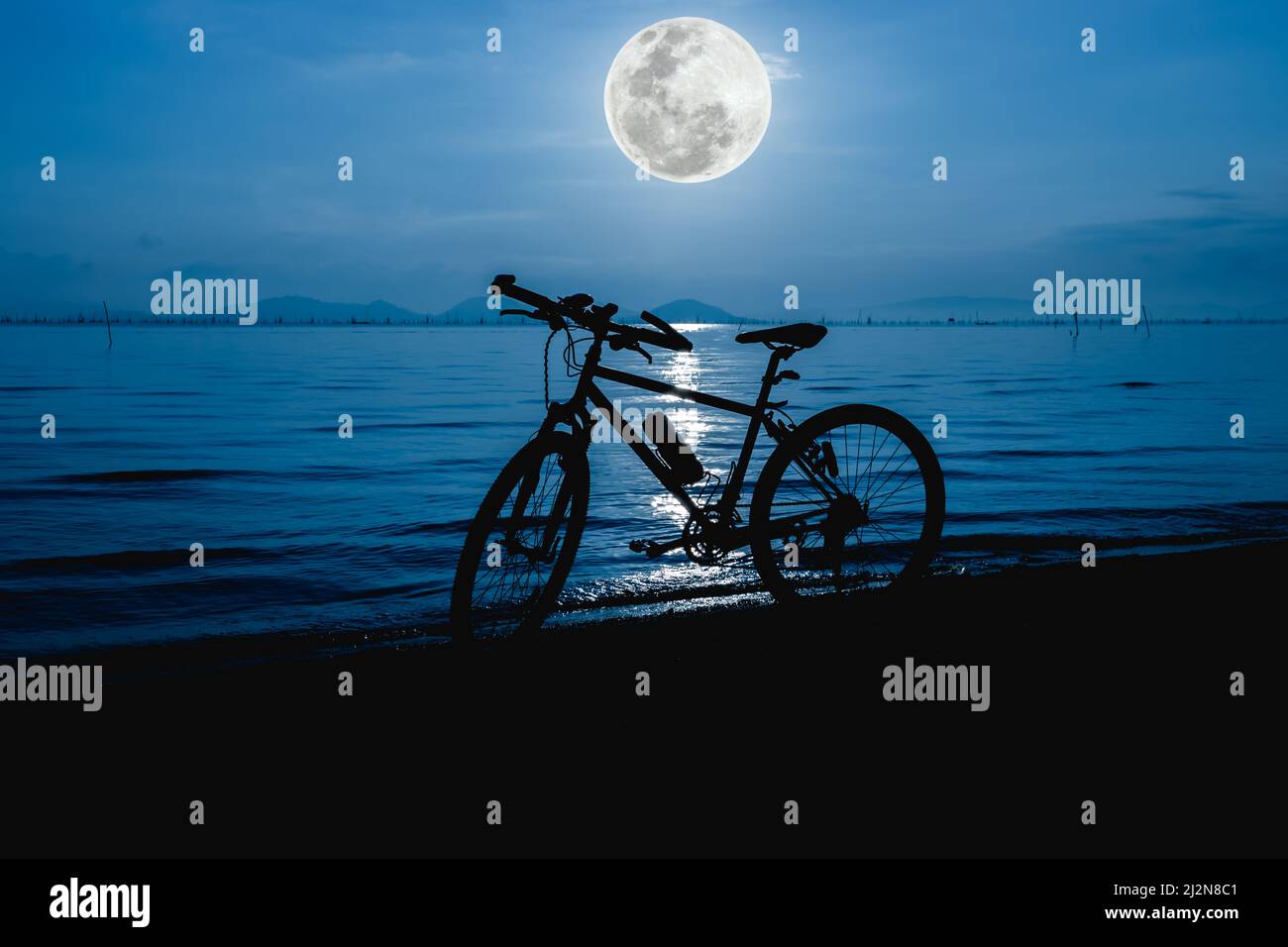 Silhouette of bicycle on the beach against beautiful full moon in the sea, blue sky background. Outdoors. The moon were NOT furnished by NASA. Stock Photo