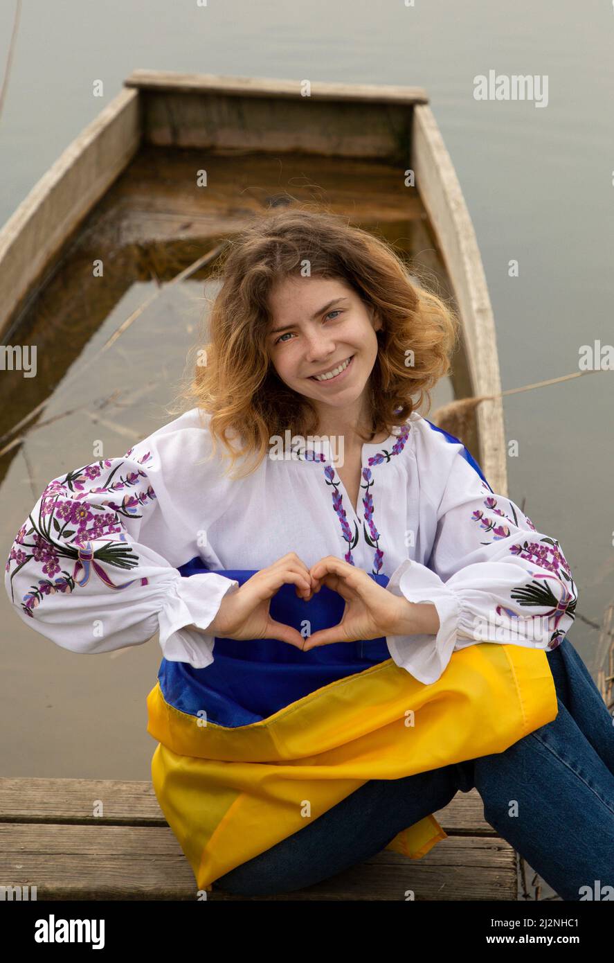 beautiful girl 17-19 years old in embroidered shirt with Ukrainian flag on river bank. hands are folded in shape of heart. Ukrainians are against war. Stock Photo