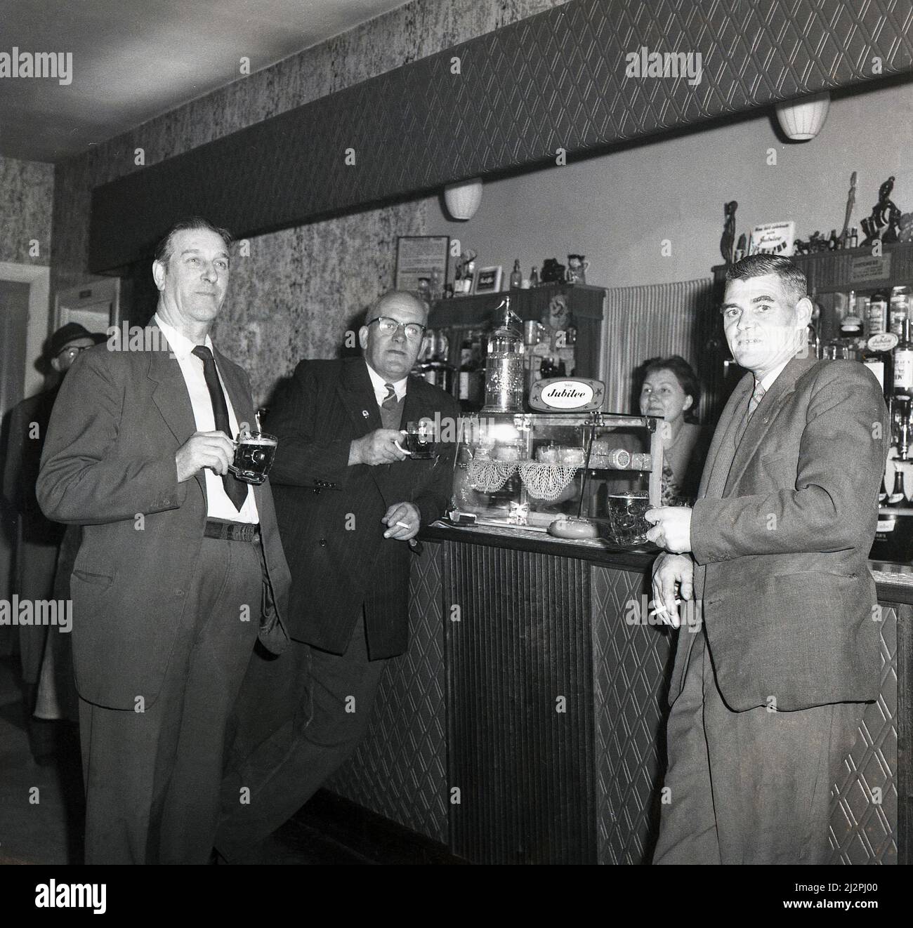 1963, historical, three me standing having a drink - and a smoke - at a bar, Stockport, Manchester, England, UK. Glass case on the bar with pork pies and sign for the Jubliee beer brand, most famously a Jubliee Stout. Jubliee beer was brewed by Hope and Anchor of Sheffield, who merged in 1962 with the Charington brewery of Mile End, London. Stock Photo