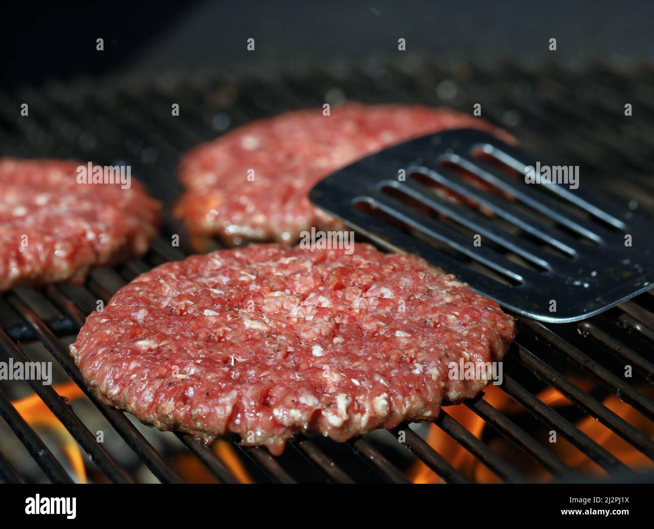 Fresh raw beef burgers on grill grate with flames and stainless steel spatula in background