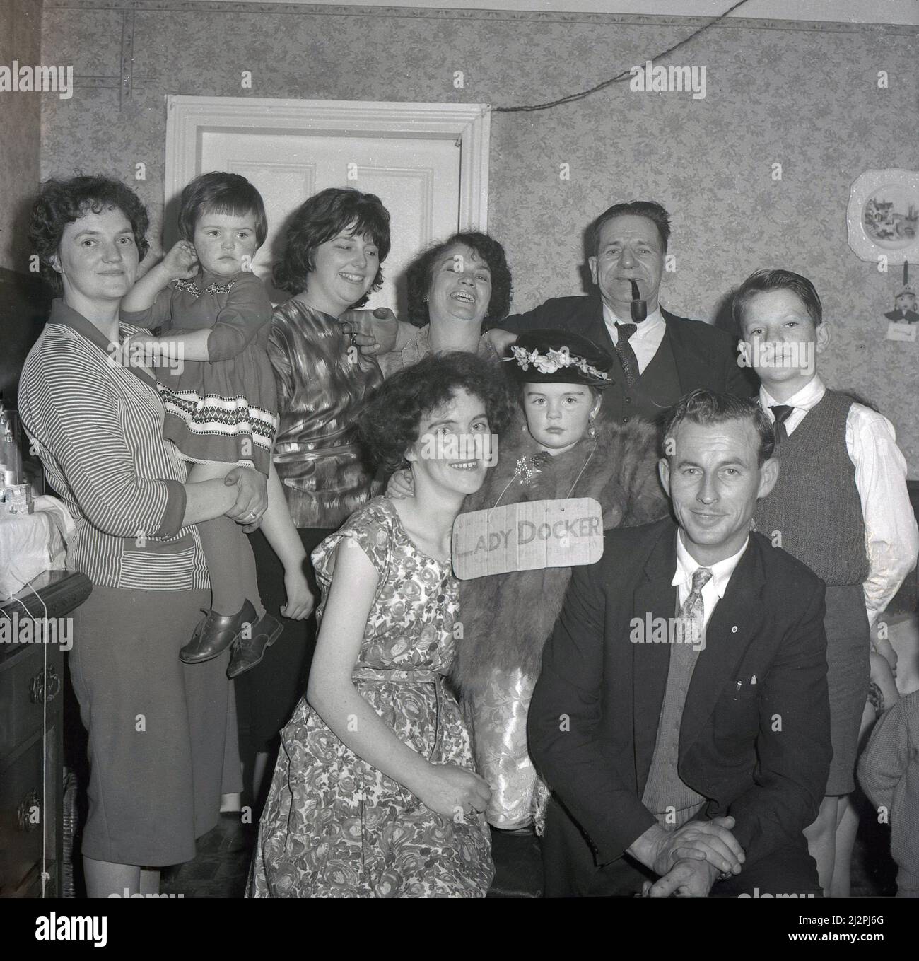 1961, historical, with her mum and dad and other family members celebrating her birthday, a young girl dressed up, as the cardboard sign on her says, as 'Lady Docker', Stockport, Manchester, England, UK. In post-war British, Lady Docker - born Norah Royce Turner in a flat over a butchers shop in Derby - was a media 'personality', a socialite. Through marriage, in particular her third marriage to Sir Bernard Docker, Chairman of the Daimler car company and a director of Midland Bank, she led a colouful, extravagant life for the time, but she never forgot her humble working class roots. Stock Photo