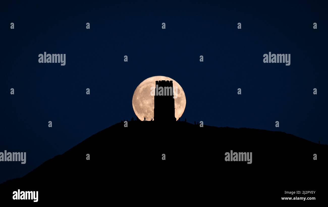 A supermoon ( Harvest Moon) rising behind Glastonbury Tor silhouetting St Michaels Tower and people on the hill. Stock Photo