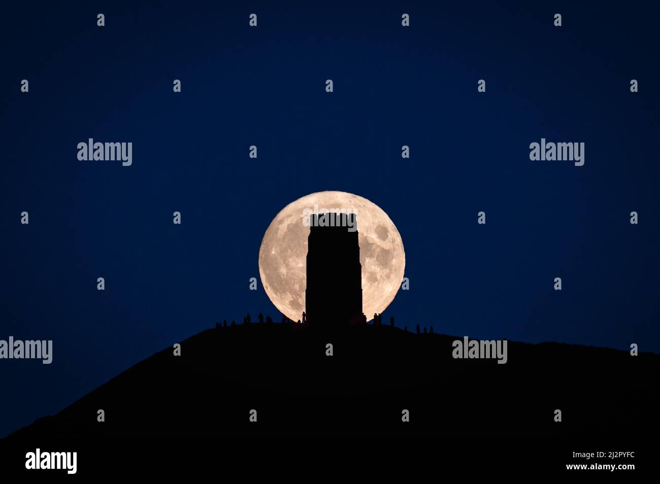 A supermoon ( Harvest Moon) rising behind Glastonbury Tor silhouetting St Michaels Tower and people on the hill. Stock Photo