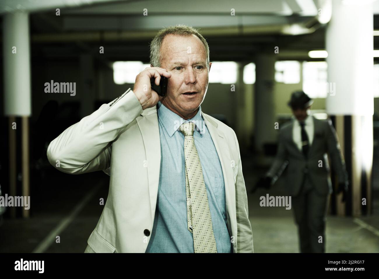 Completely unaware his life is about to change. An oblivious businessman talking on his cellphone as an armed assailant approaches from the shadows in Stock Photo