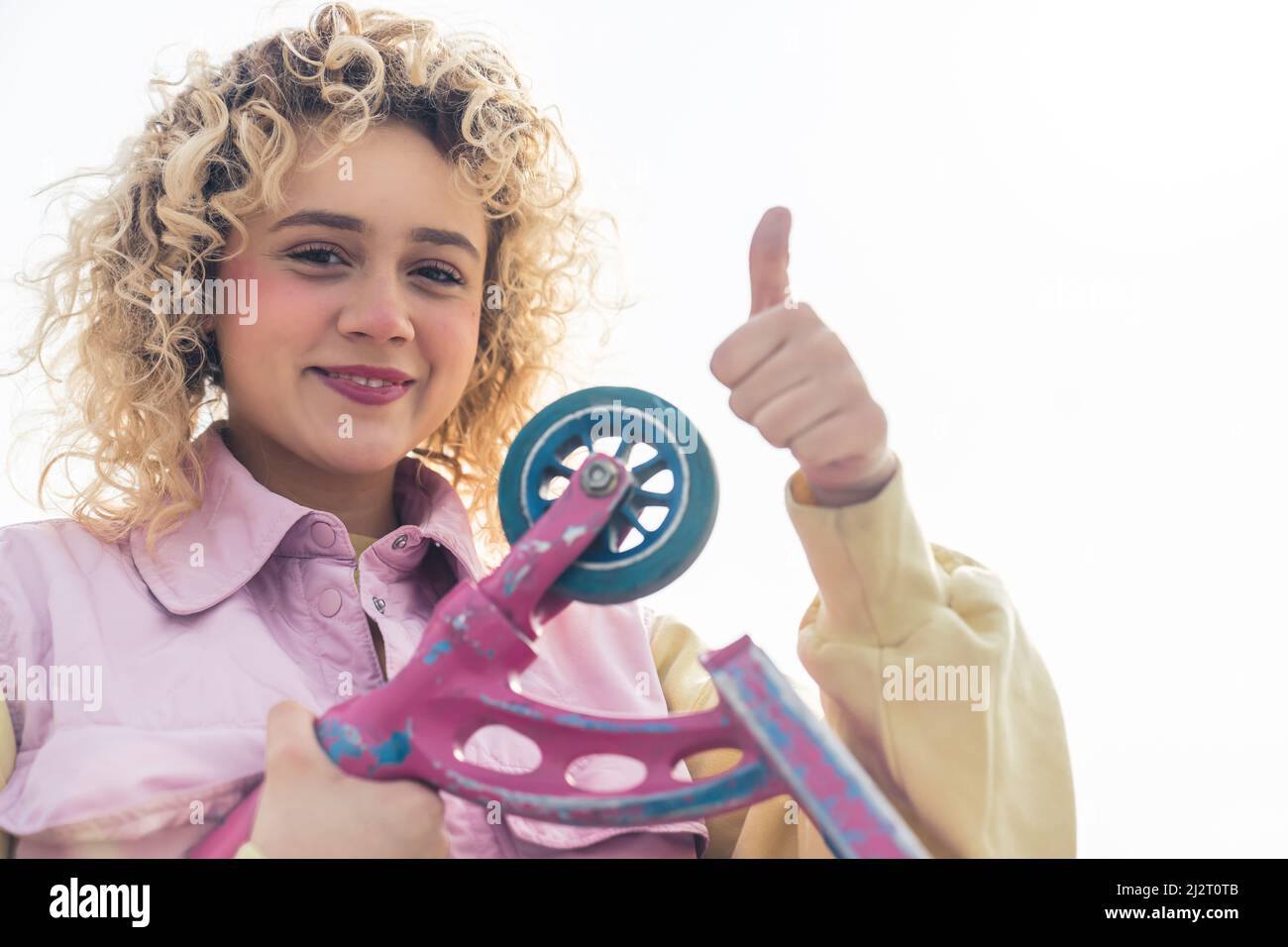 Beautiful young blond hipster woman looking at the camera, showing thumb up and holding a scooter, medium closeup copy space white background . High quality photo Stock Photo