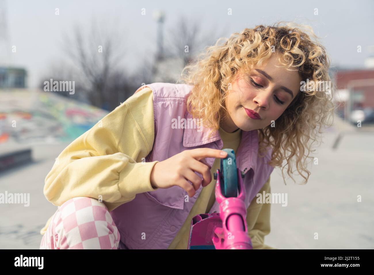 Young blond curly haired woman posing with a pink scooter, looking down and touching the wheel, portrait copy space sports ground background . High quality photo Stock Photo