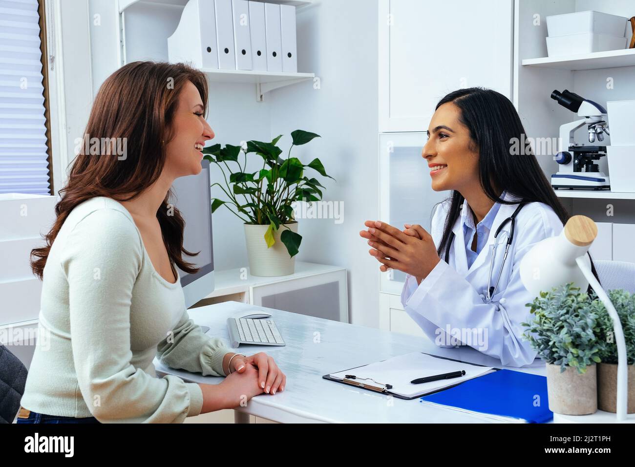 Laughing female doctor and lady client patient discussing something while sitting at the table in medical center healthcare industry Stock Photo