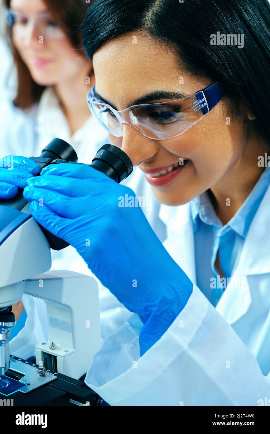 Young female scientist using microscope while her colleague working in background healthcare industry Stock Photo