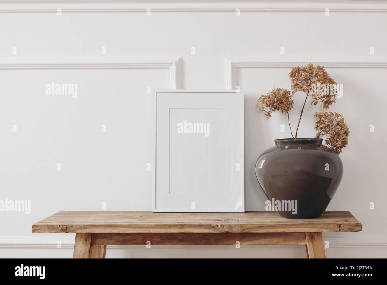 Brown bold vase. Dry hydrangea flowers on old wooden bench. Blank black picture frame mockup. White wall moulding background, stucco decor. Empty copy Stock Photo
