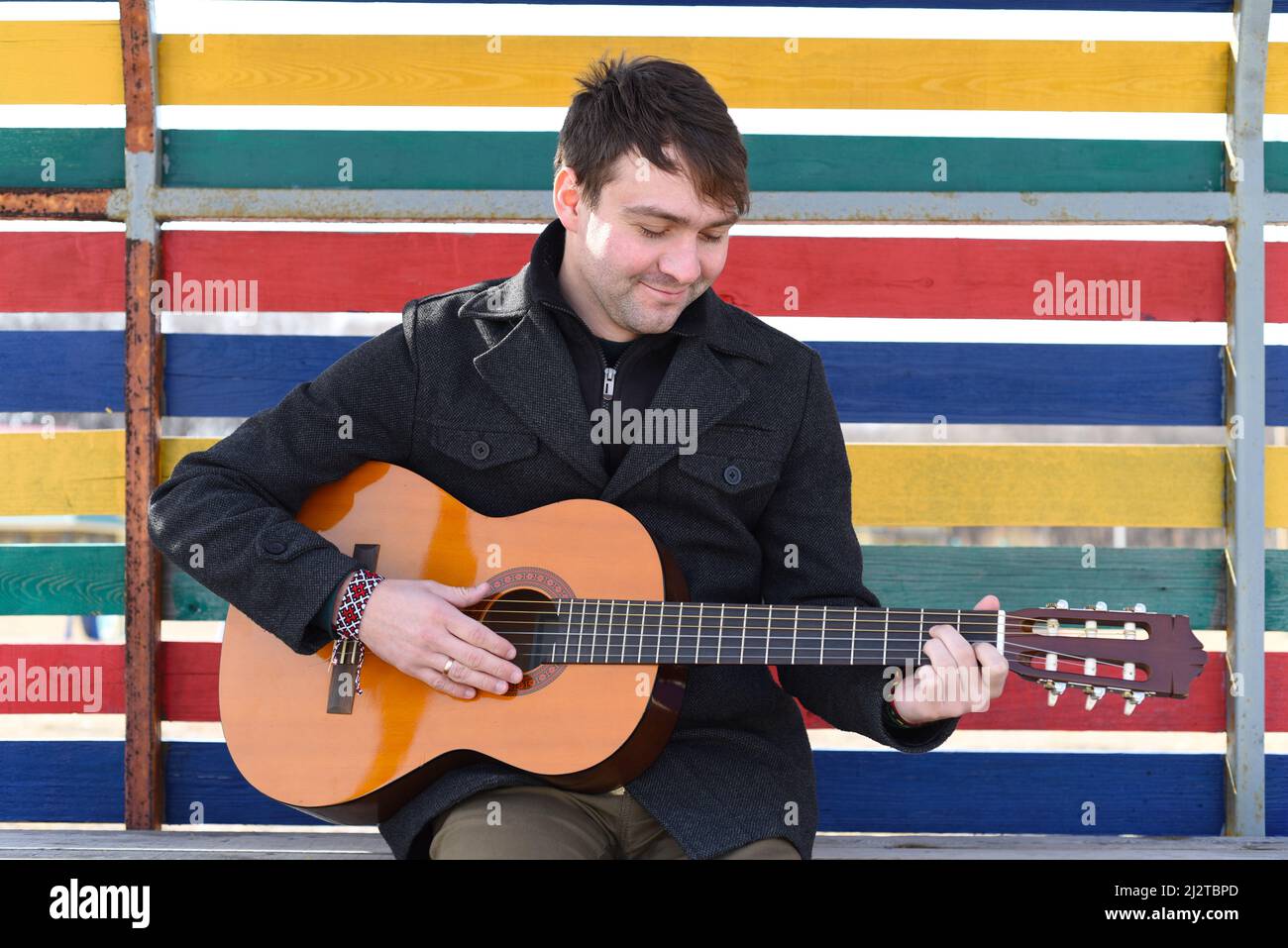 Smiling young man playing guitar and sitting against colorful wall Stock Photo