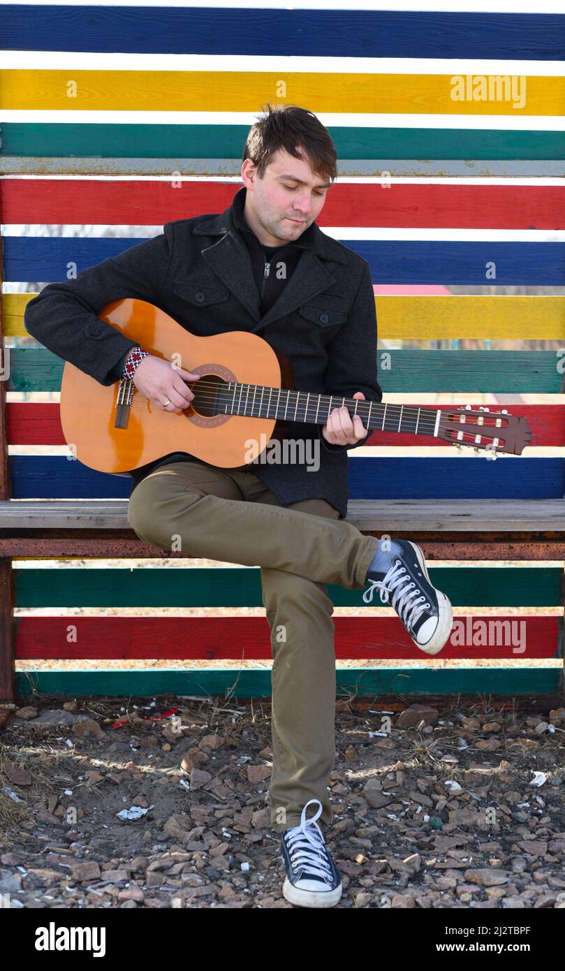 Smiling young man playing guitar and sitting against colorful wall Stock Photo