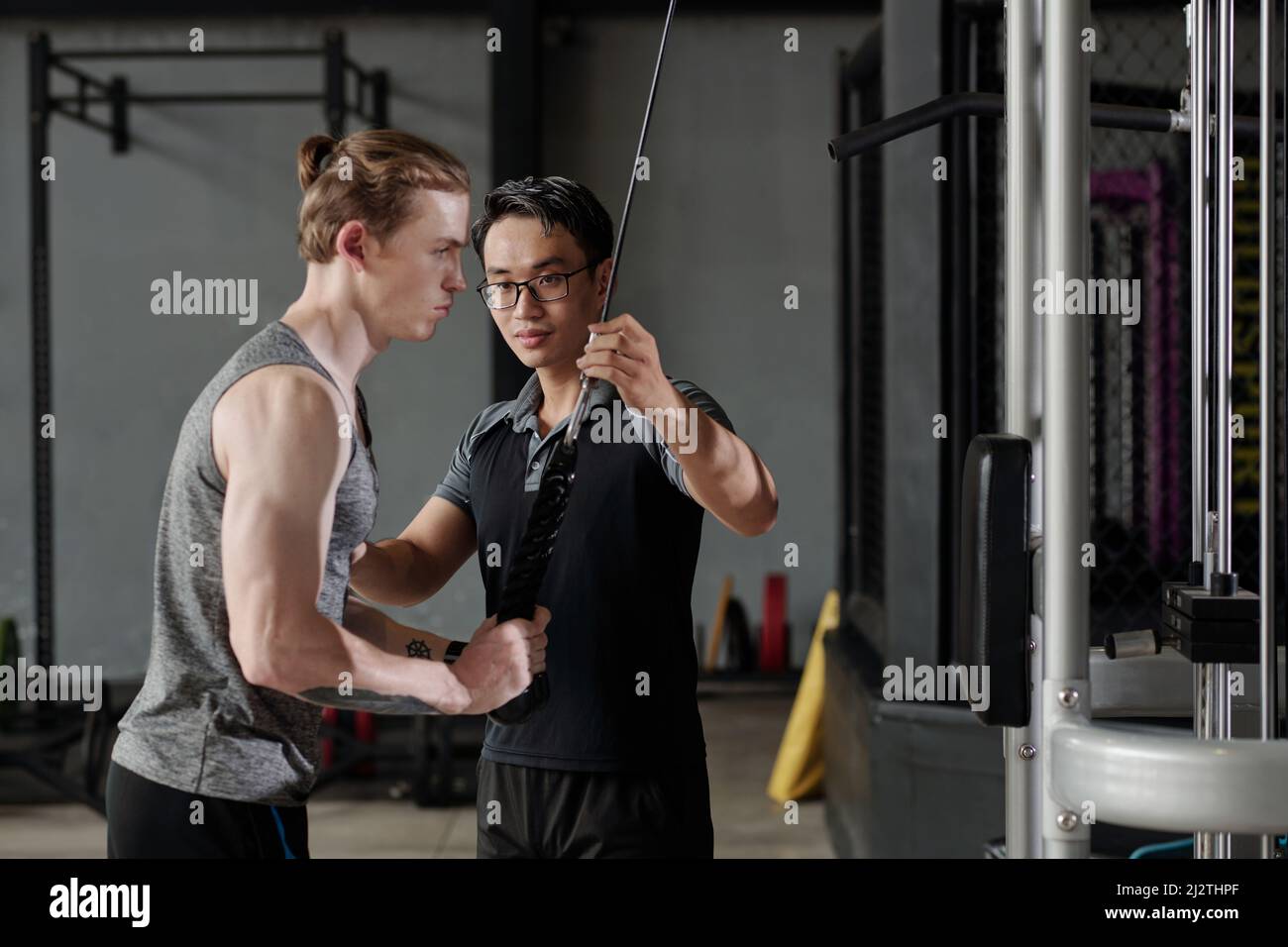 Fitness trainer correcting muscular young man doing cable pull down