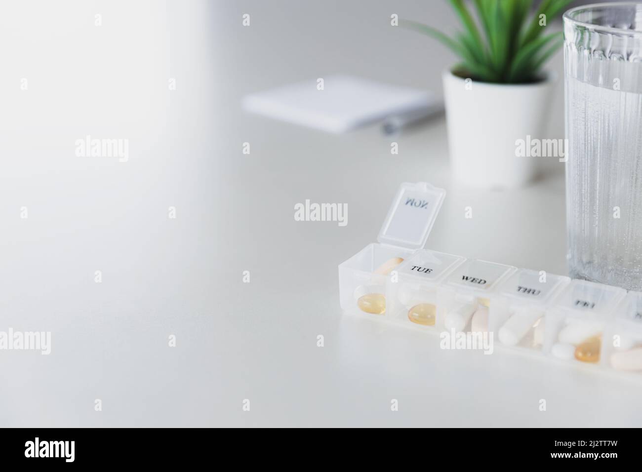 Closeup of medical pill box with doses of tablets for daily take a medicine with white, yellow drugs and capsules used for treatment, cure the disease. Glass cup of water on table, gray background. Stock Photo
