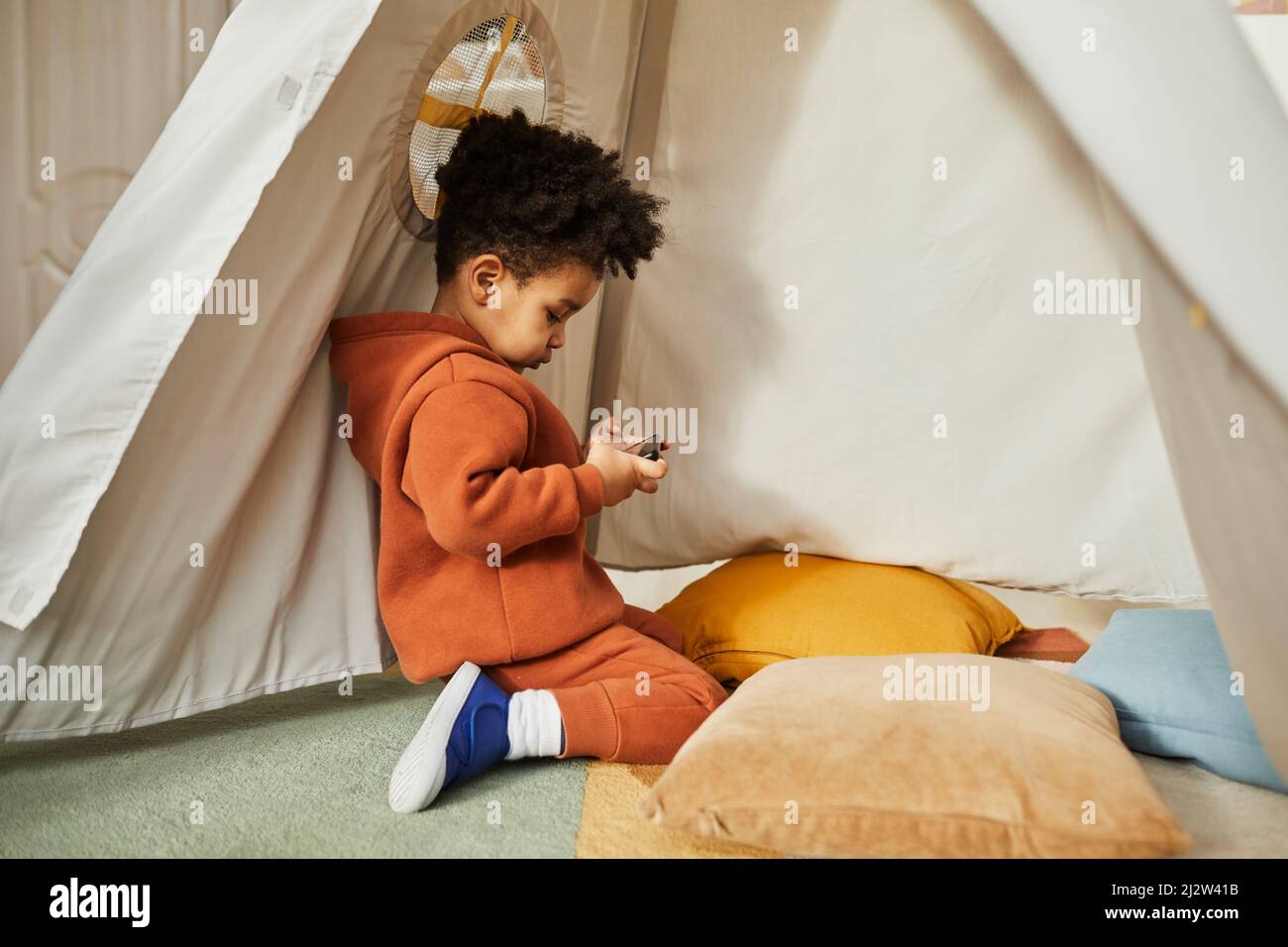 Minimal side view portrait of little African American boy using smartphone in play tent and wearing trendy outfit, gen Alpha, copy space Stock Photo