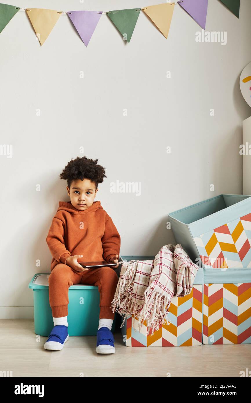 Minimal full length portrait of black toddler boy holding tablet and looking at camera in cute kids room interior Stock Photo