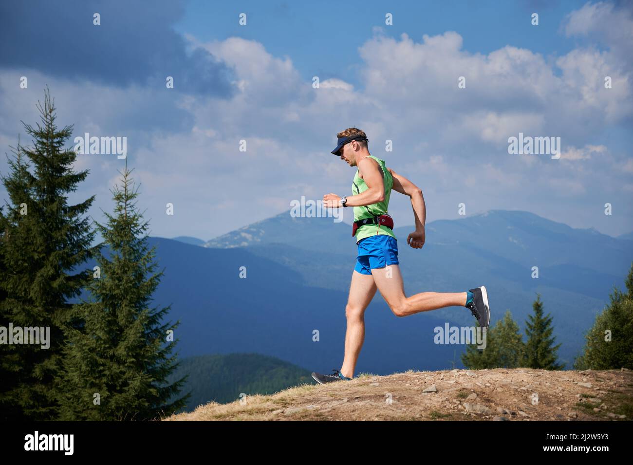 Light-skinned male runner of trail with sports bag on his waist and cap running from stone top of mountain hill. Healthy man jogging downhill among the summer nice mountain scenery. Stock Photo