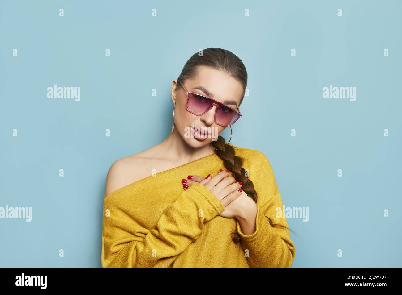 Lovely young woman presses palms to heart, feels touched and grateful, dressed in yellow jumper, isolated over blue background Stock Photo